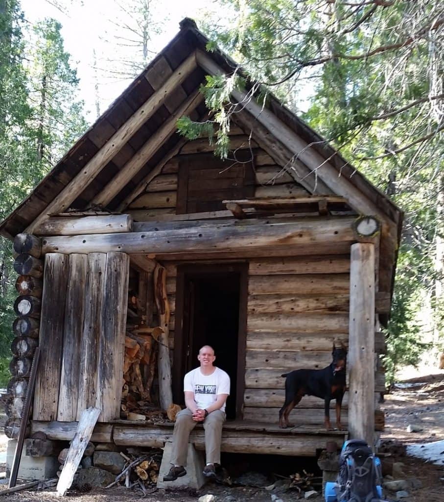 Man en zijn Doberman zitten op de veranda van een rustieke hut.
